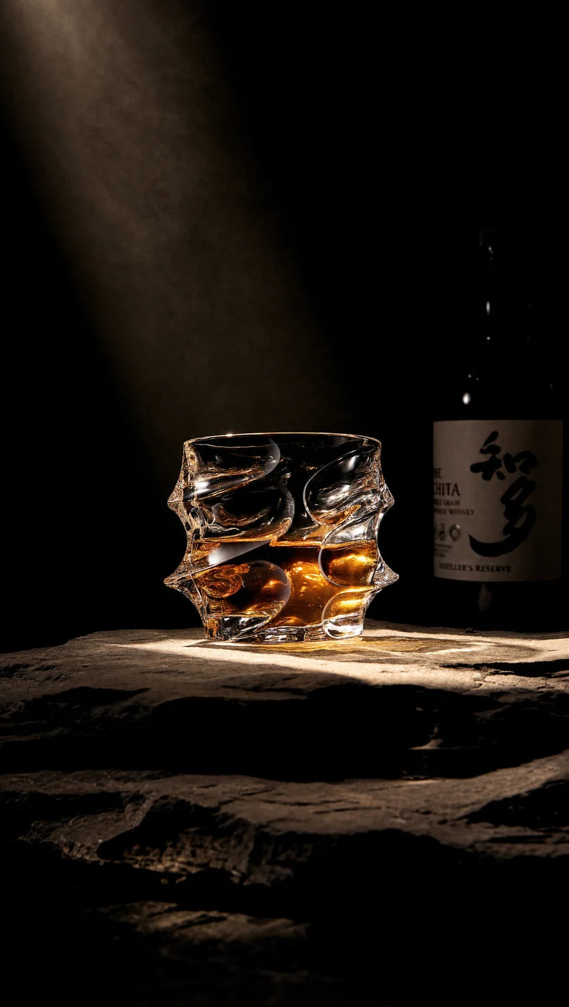 A uniquely shaped glass of whisky sits on a stone surface, dramatically lit, with a bottle behind it.