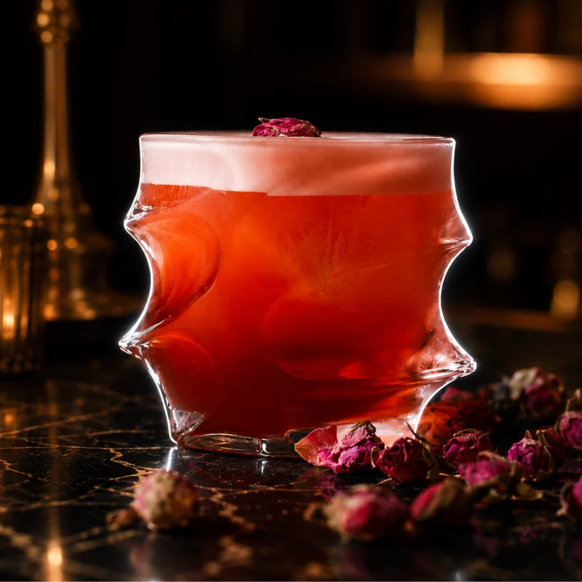 A red foamy cocktail in a unique, spiky glass sits on a marble counter surrounded by dried rosebuds.